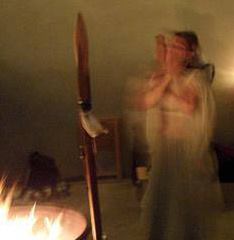 Ren Zatopek stands behind a firepit and a wooden spear wearing something long, sheer, and white. She is blurry from movement, her hands covering her face. The blur of the movement alludes to the feeling of the trance in this ritual.