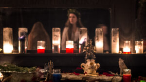 A beautiful altar is laid with an animal skull, a bronze statue of Pan, a ritual bell and wand, and many lit candles. Behind the altar a large mirror reflects the candlelight and a few figures in the darkness of the ritual space. In the center one of the figures is almost recognizable as Ren Zatopek, perhaps with a crown of laurel on her hair, which hangs long and loose. She wears something sheer and white. There is a feeling of trance in this image from the way the figures are blurred.