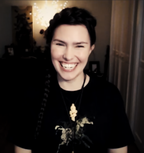 A clip of Ren Zatopek from a Hedge + Riders online class, her dark hair in a braid that goes up over her head and down her side, a big smile on her rosy face, wearing a black t-shirt with a gold print of a horse on it.