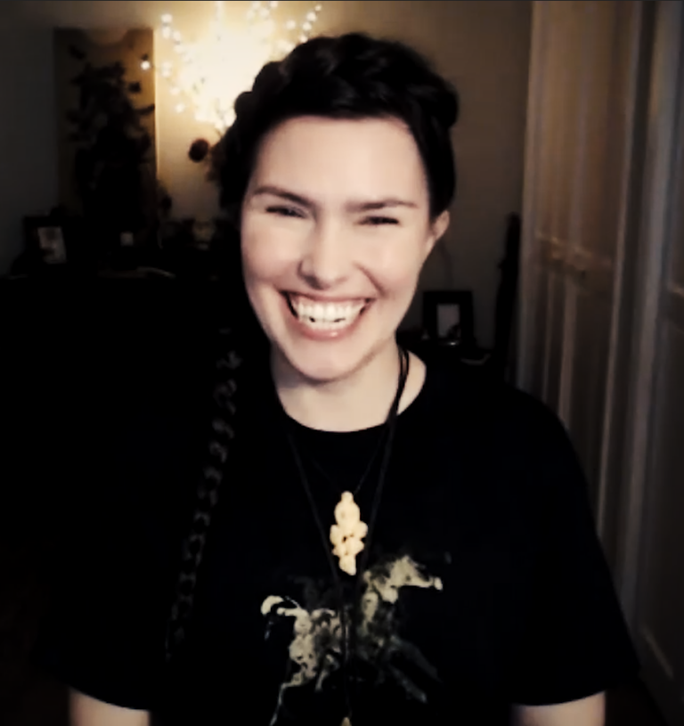 A clip of Ren Zatopek from a Hedge + Riders online class, her dark hair in a braid that goes up over her head and down her side, a big smile on her rosy face, wearing a black t-shirt with a gold print of a horse on it.