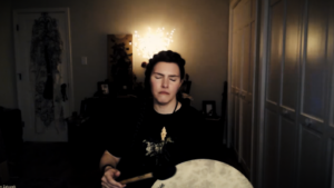 A clip of Ren Zatopek from a Hedge + Riders online class, her eyes are closed as she holds a frame drum and a beater stick with a black felt wrapping on the end. She wears a black shirt with a gold horse printed on it and a necklace with a skull pendant that appears to be made of bone.