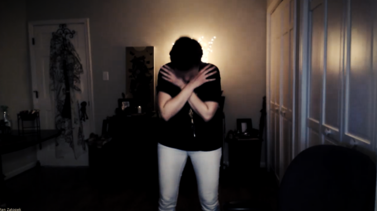 A clip of Ren Zatopek from a Hedge + Riders online class, she stands with her head bowed and her arms crossed, as if hugging herself. The room is dark save a glowing vase in the distance.