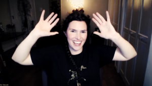 A clip of Ren Zatopek from a Hedge + Riders online class, her dark hair in a braid that goes up over her head and down her side, a big smile on her rosy face, wearing a black t-shirt with a gold print of a horse on it. She's got both hands up, gesturing enthusiastically.