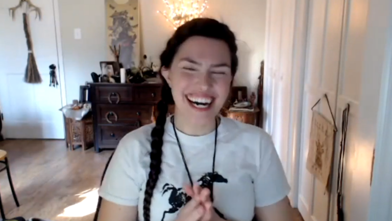 A clip of Ren Zatopek from a Hedge + Riders online class, unleashing a huge, laughing smile. The room is bright and an ornate altar is just visible on the wall behind her. Her dark hair is braided. She wears a white t-shirt with a black horse painted on it.
