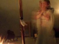 Ren Zatopek stands behind a firepit and a wooden spear wearing something long, sheer, and white. She is blurry from movement, her hands covering her face. The blur of the movement alludes to the feeling of the trance in this ritual.