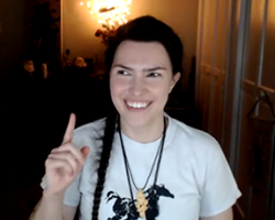 A clip of Ren Zatopek from a Hedge + Riders online class. She has a mischievous smile and holds up one finger—she's about to make a point! Twinkle lights on the altar behind her illuminate the dark room. Her white t-shirt with painted black horse is bright in the foreground.